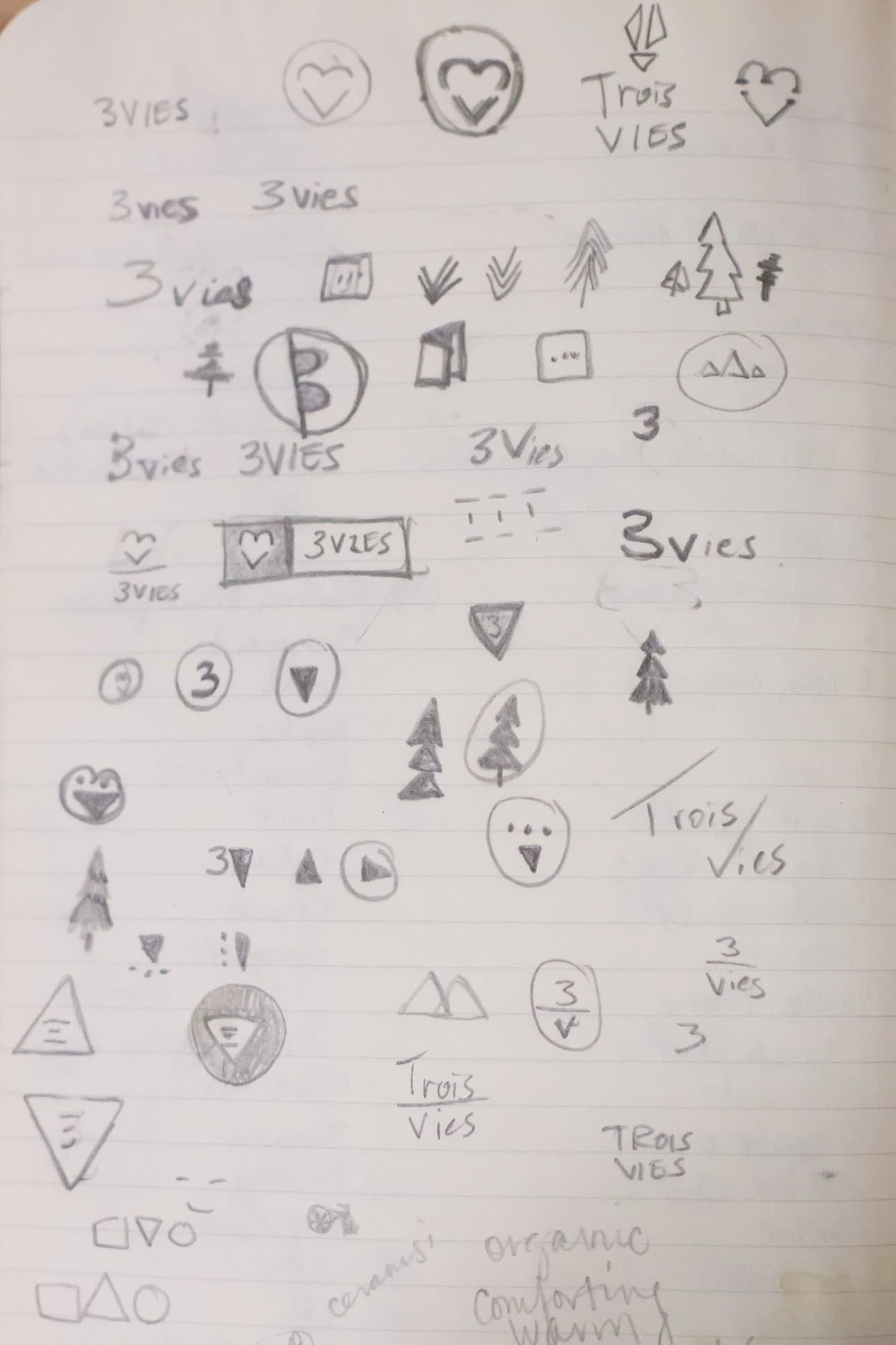 Notebook page of pencil logo sketches exploring 3vies and Trois Vies wordmarks, trees, hearts and triangles.