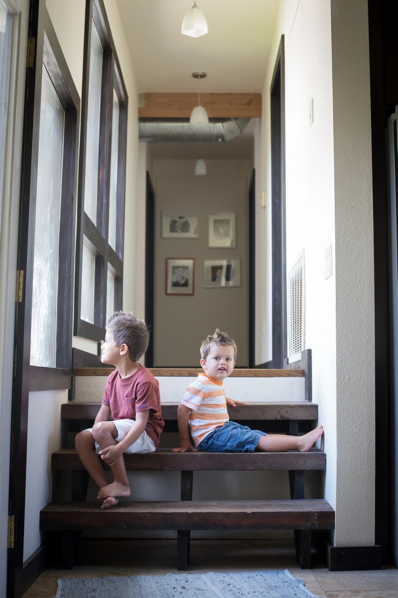 Two young brothers sit on wooden stairs in the home's bright hallway lined with framed photos.