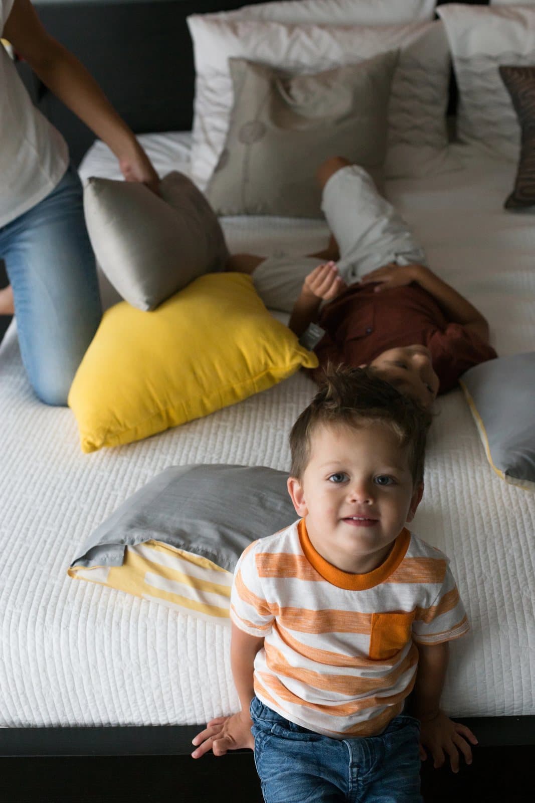 A toddler in an orange striped shirt peers up from the edge of the bed mid pillow fight.