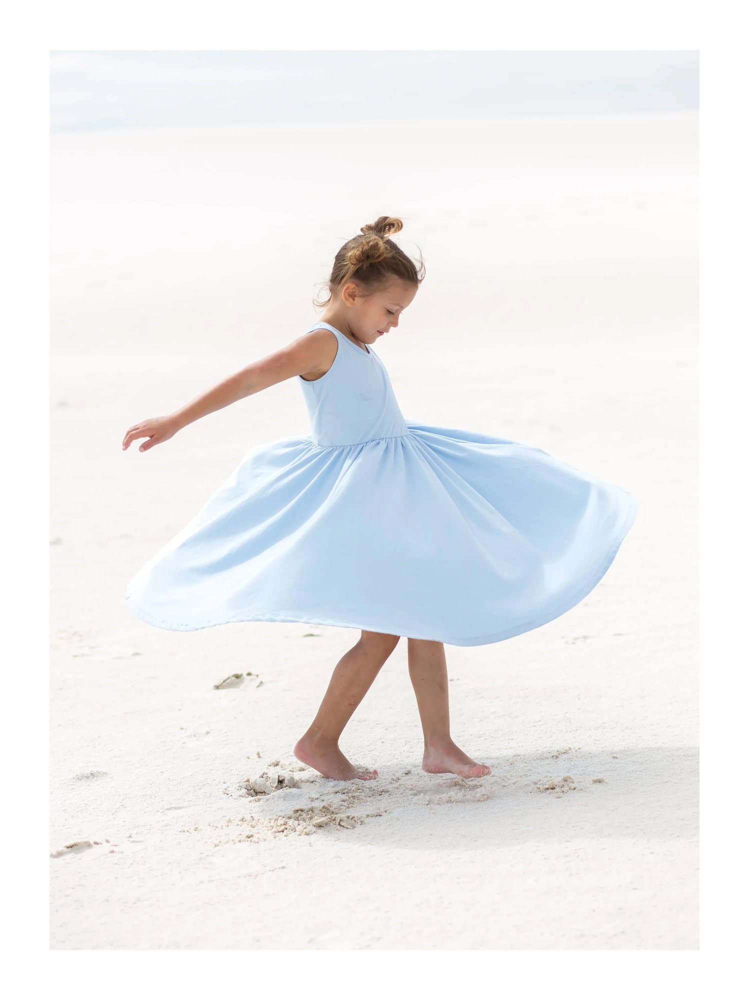 Aila twirling barefoot in a light blue dress on white sand, TIMELESS print Aila.