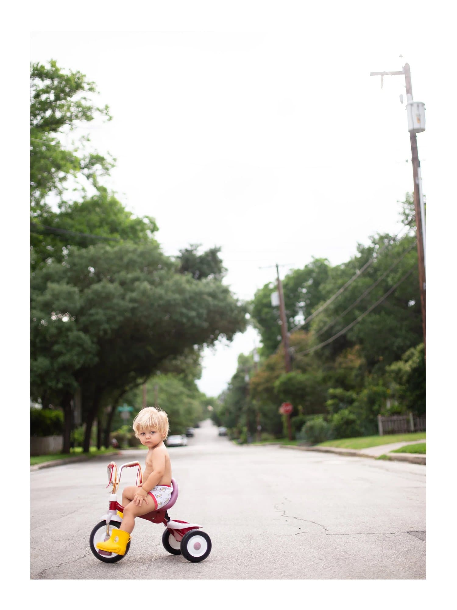 Luca in rain boots on a red tricycle in a tree-lined street, TIMELESS print Luca.