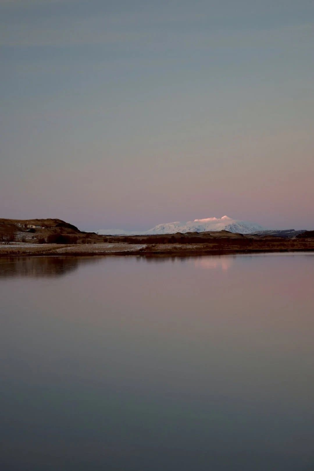 A calm lake at dusk reflecting a pink sky, with a snowcapped mountain on the horizon.