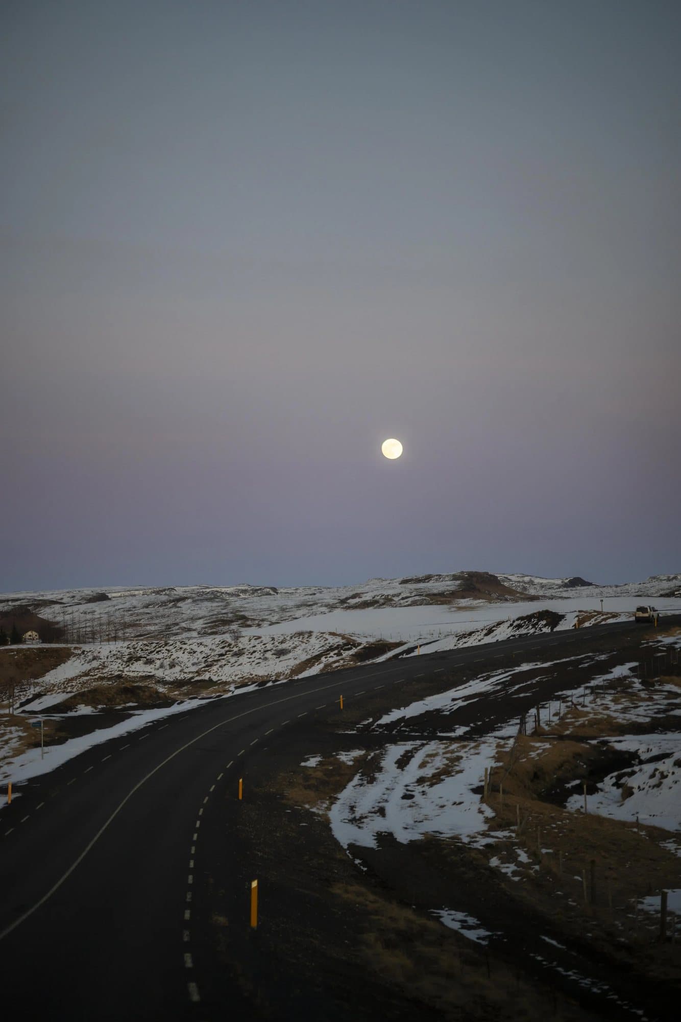 A full moon over a winding road through snow-patched Icelandic hills at dusk.