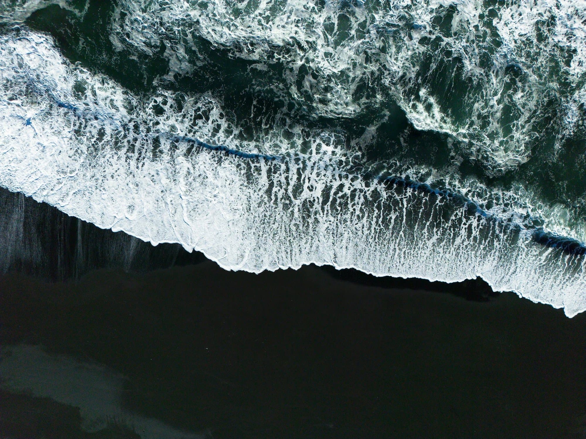 An aerial view of white surf breaking onto a black sand beach in Iceland.