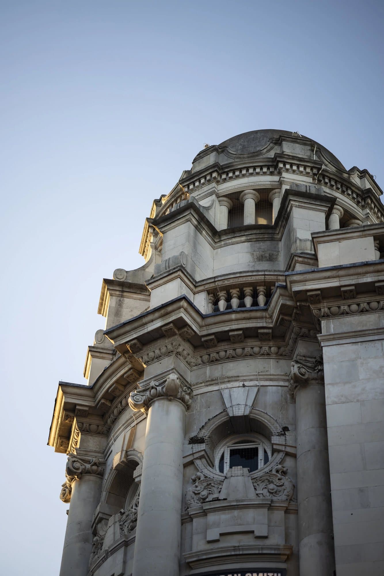 Looking up at an ornate domed stone building corner in London against a pale blue sky.