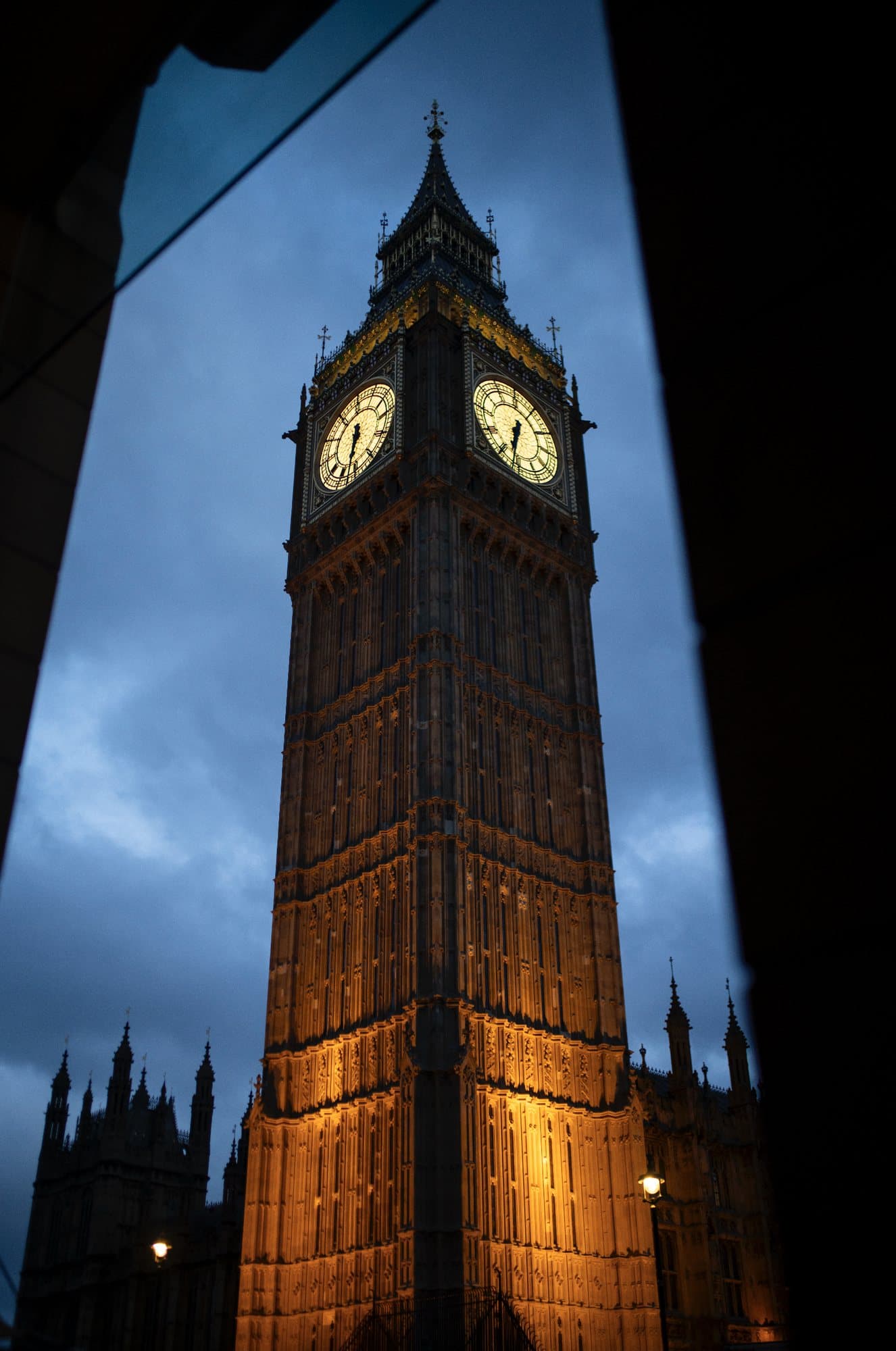 Big Ben lit warm gold against a deep blue evening sky, framed by dark foreground shapes.