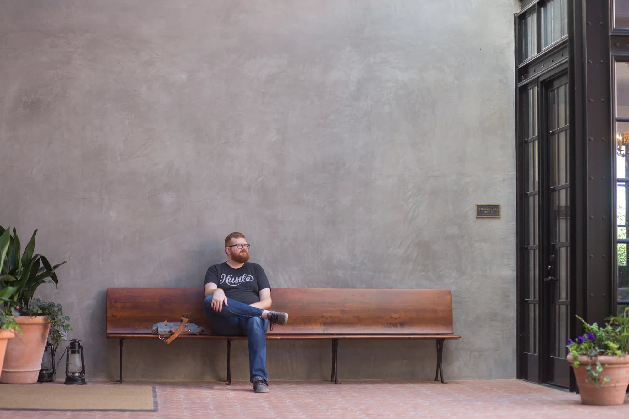 Kyle Adams sits on a wooden bench against a wide plaster wall between potted plants.