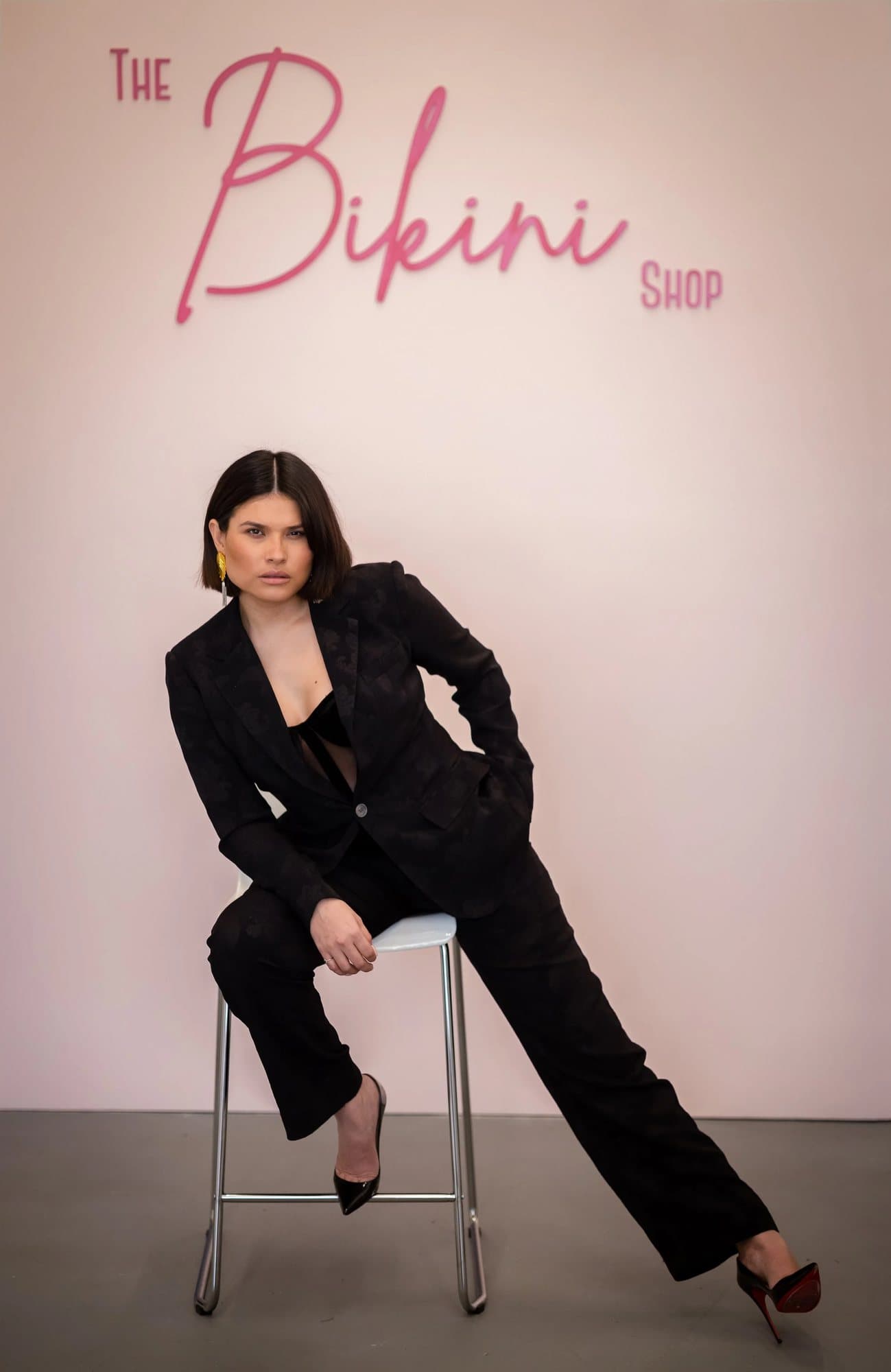 Grecia Valencia, featured in Volume 37, in a black suit on a stool beneath The Bikini Shop sign.