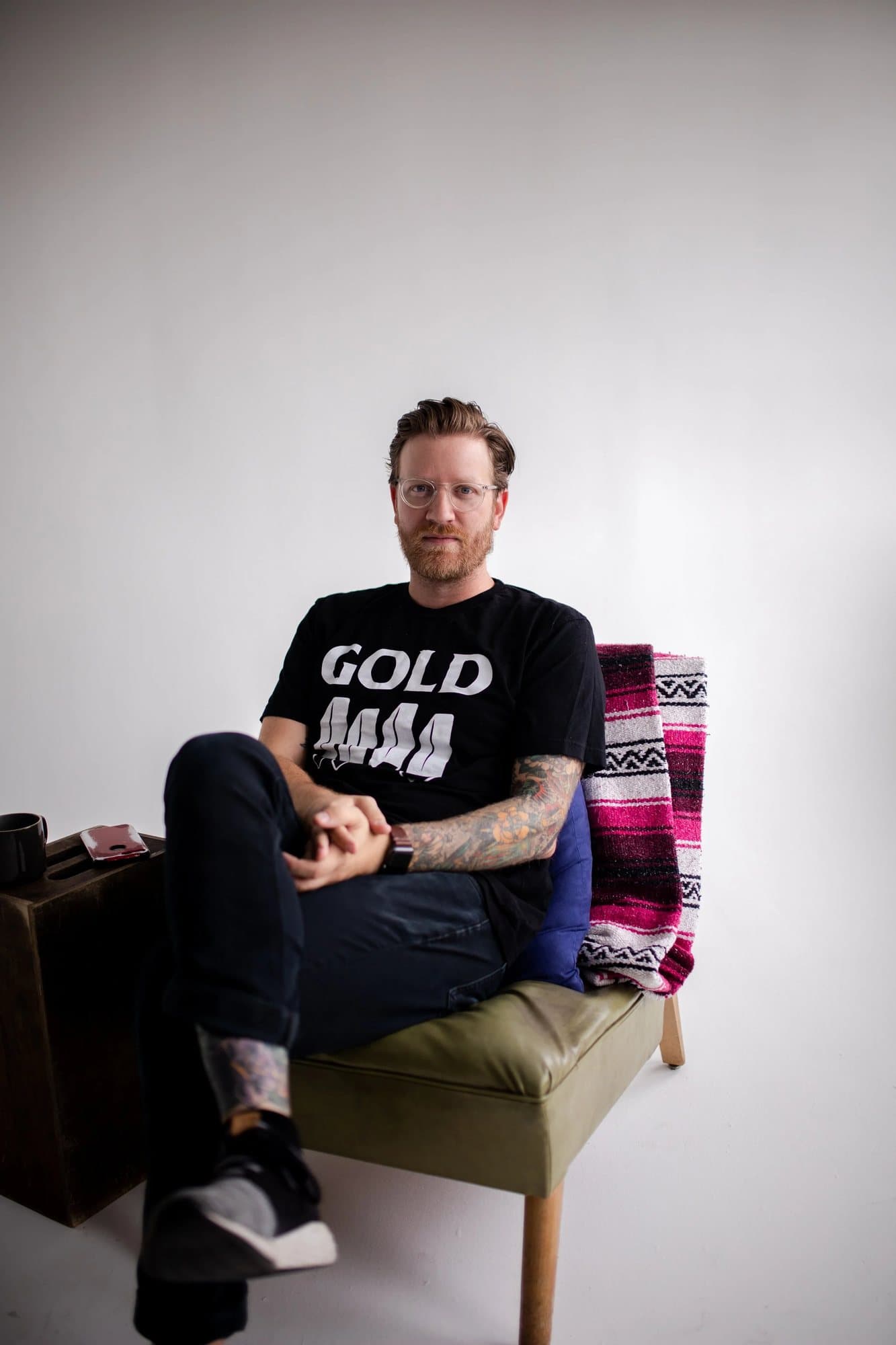 Josh Huskin, featured in Volume 34, seated in a GOLD t-shirt on a green chair in a studio.