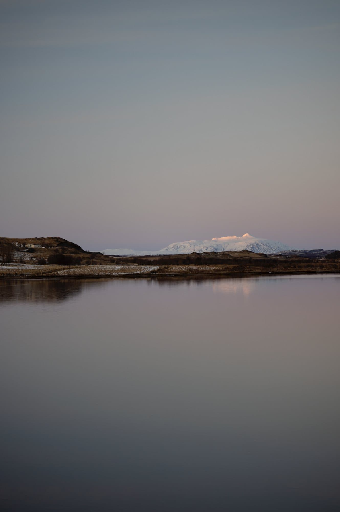 A calm lake reflecting a distant snowy mountain at dusk in Iceland.