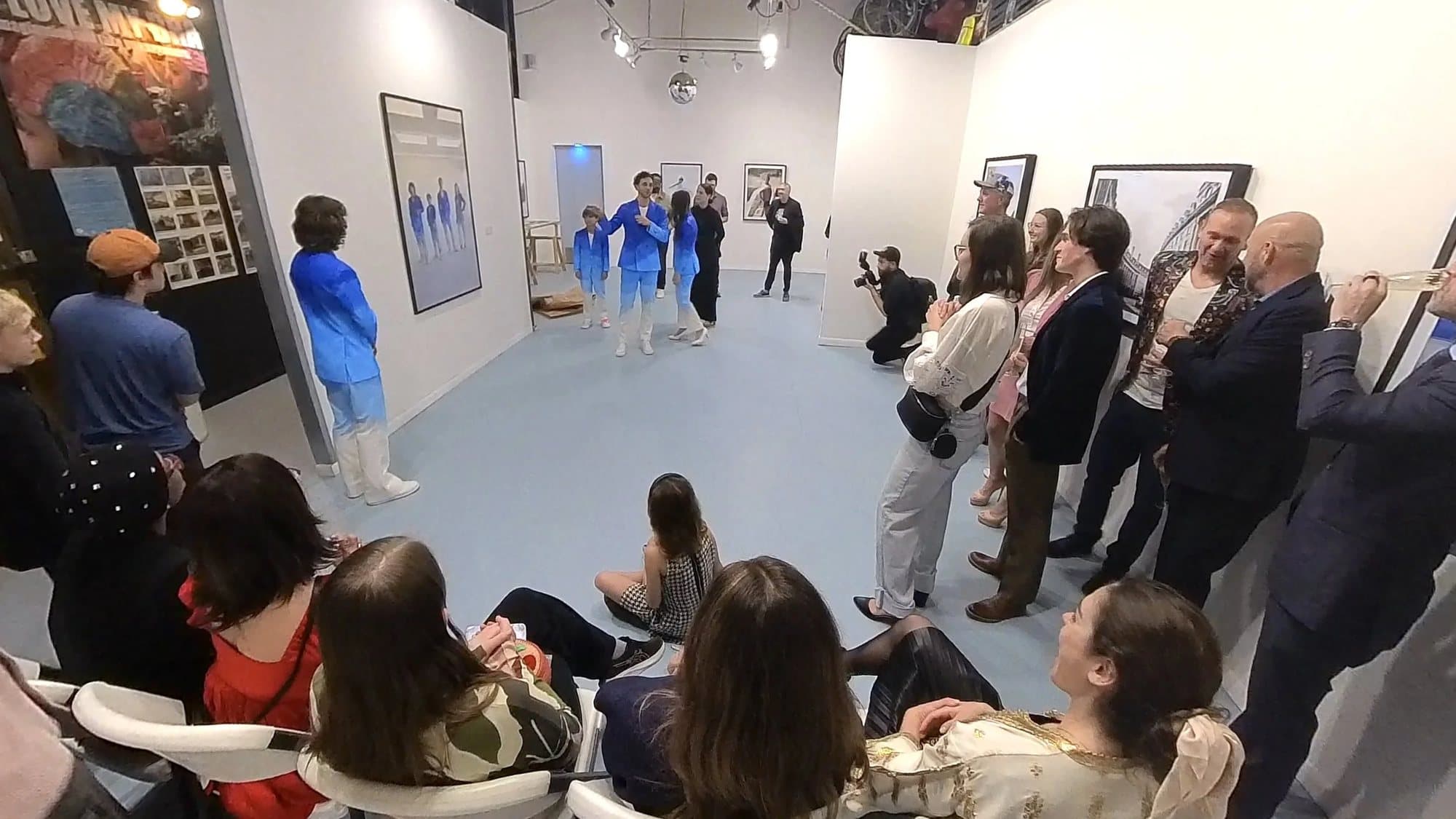 Opening night crowd at 1906 Gallery watching models in blue painted Timeless suits walk between framed prints.