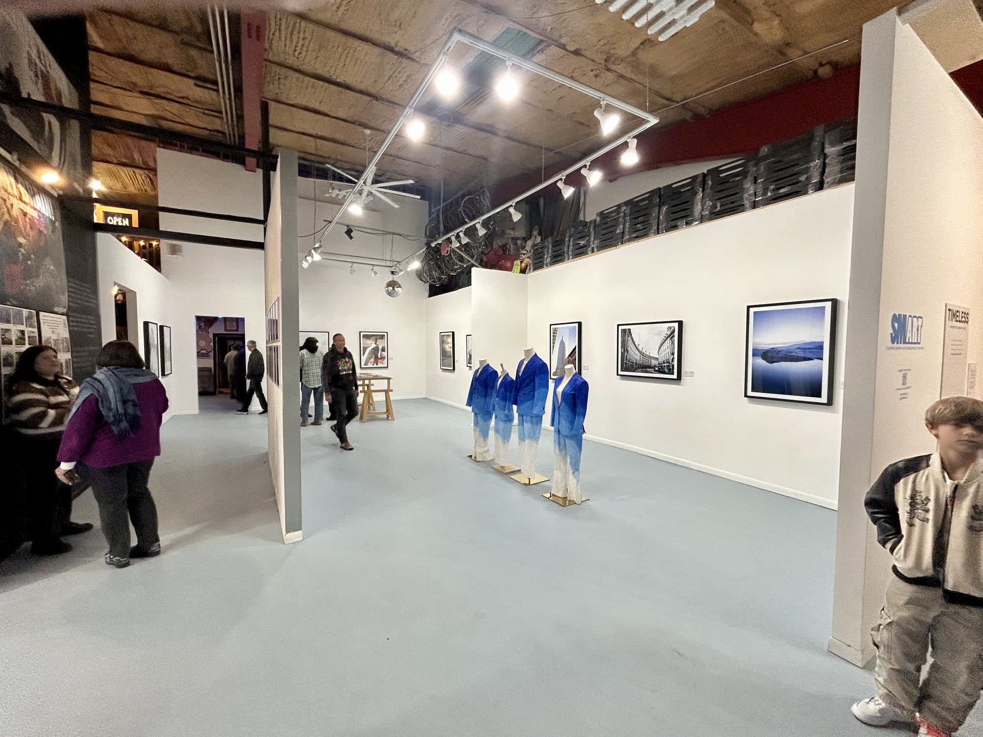 The TIMELESS exhibition: four hand-painted blue suits on stands in a white gallery, with visitors and framed photographs