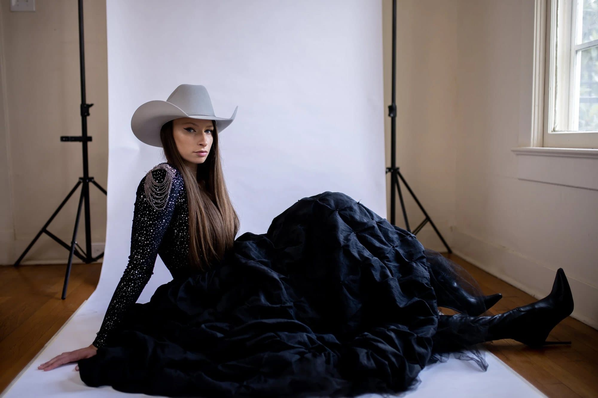 Saige Thomas in a cowboy hat and full black skirt, seated on white backdrop paper in a studio.