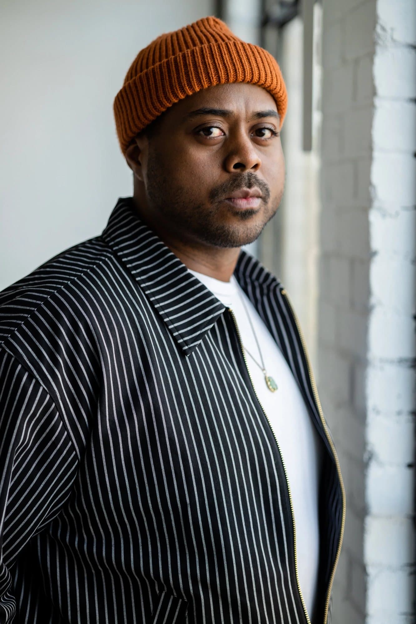 Berto Boushae in an orange beanie and pinstriped jacket beside a white brick wall.