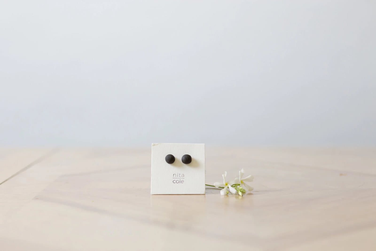 A small card of dark stud earrings rests on a pale wood floor beside a sprig of flowers.
