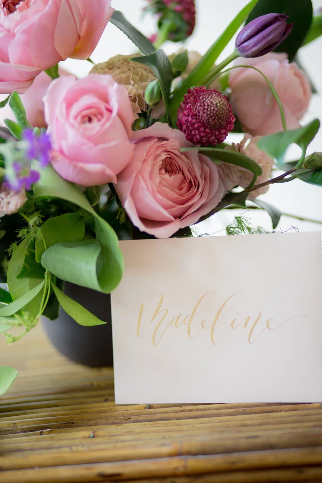 Pink roses and purple blooms above a place card hand-lettered Madeline in gold calligraphy.