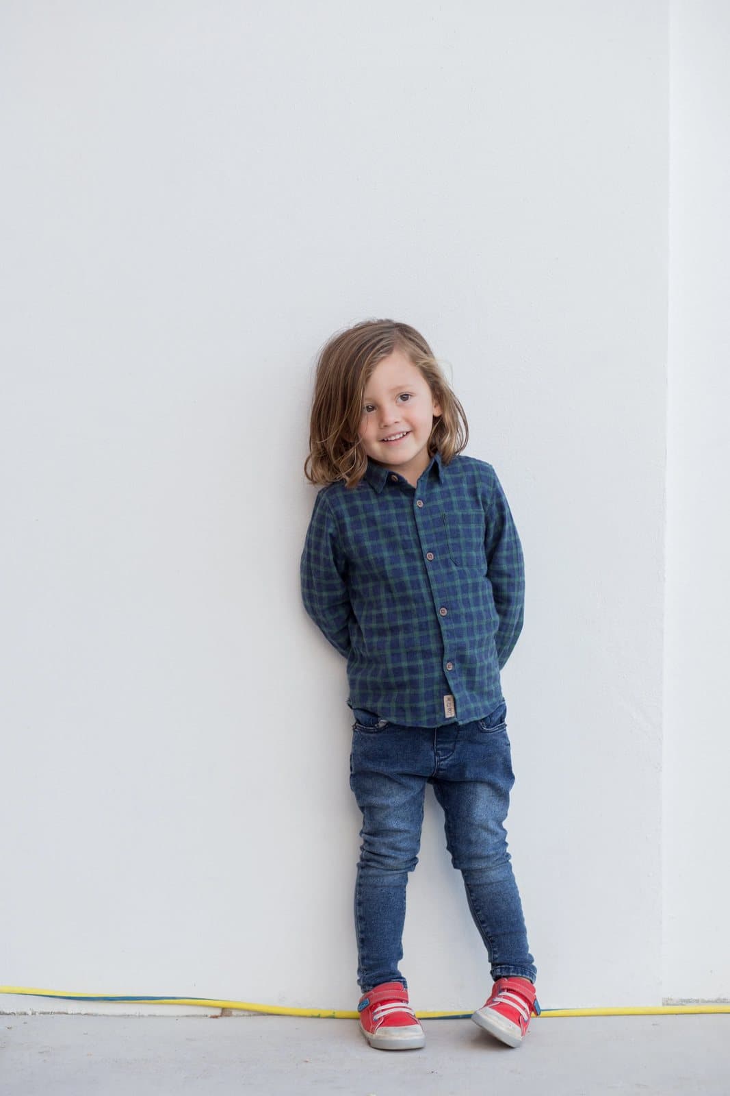 A smiling young boy in a plaid shirt and red sneakers leans against a white wall.