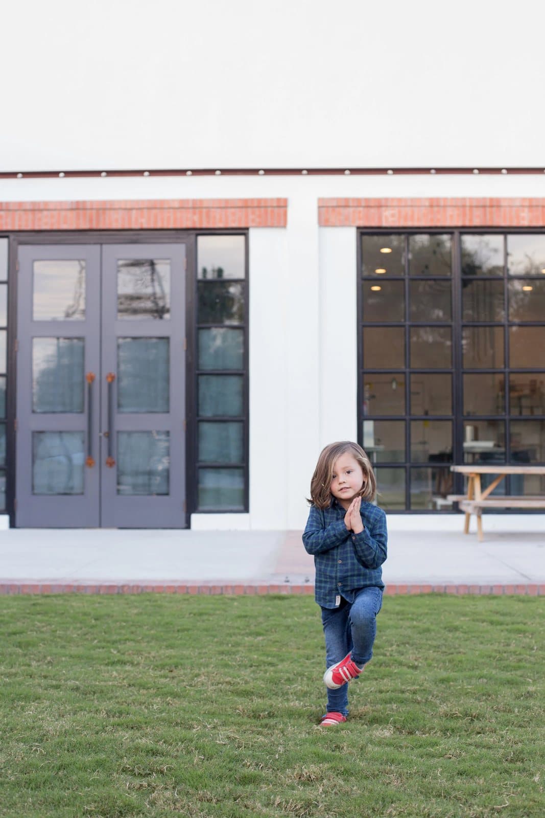 A small boy in plaid poses playfully on a lawn in front of a white building with dark doors.