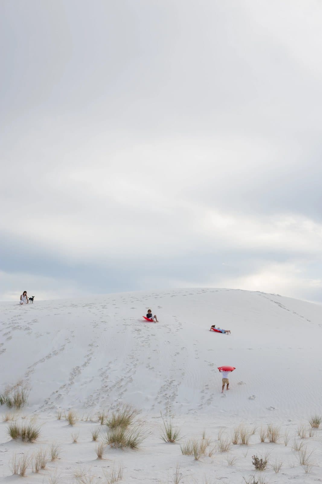 Children in bright clothes play on a vast white sand dune under an overcast sky.