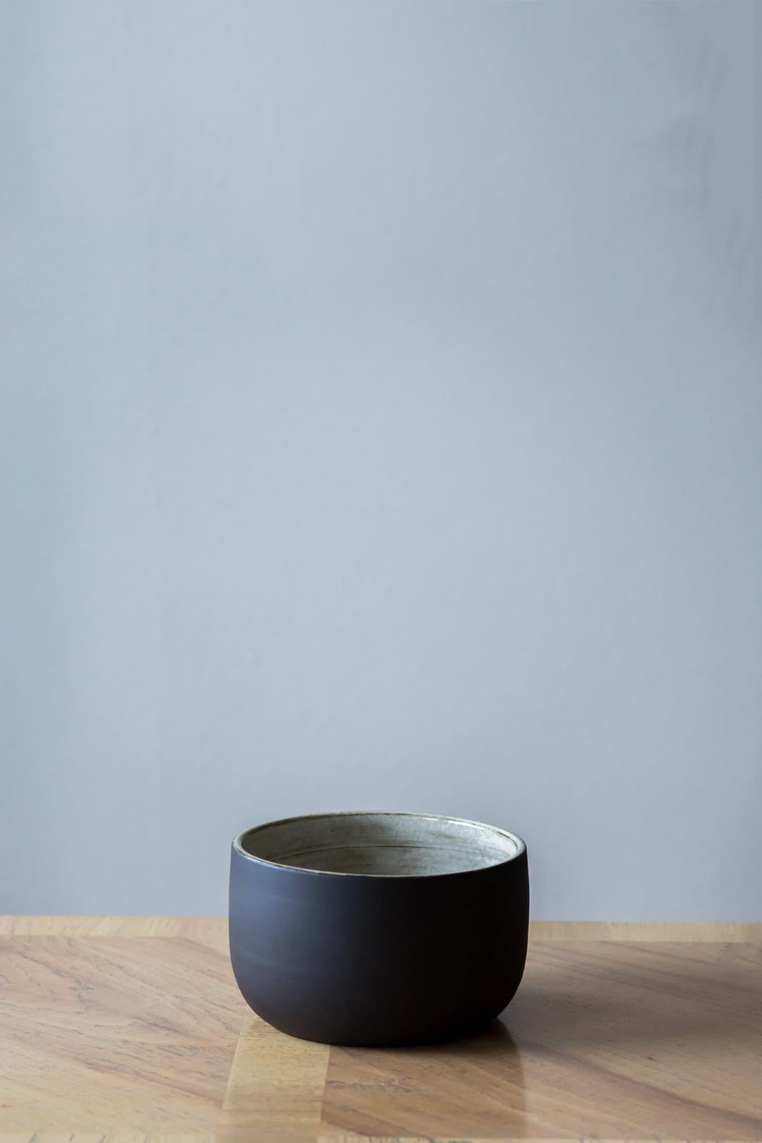 A small matte black ceramic bowl on a wood table against a pale blue wall, product photography.