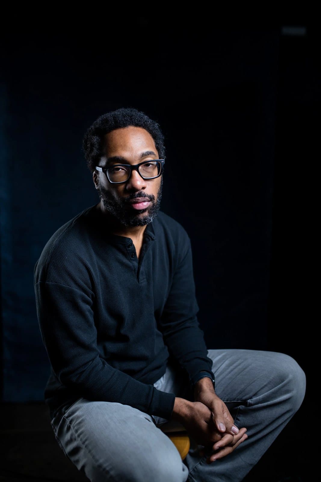 A bearded man in glasses and a black sweater sits against a dark studio background.
