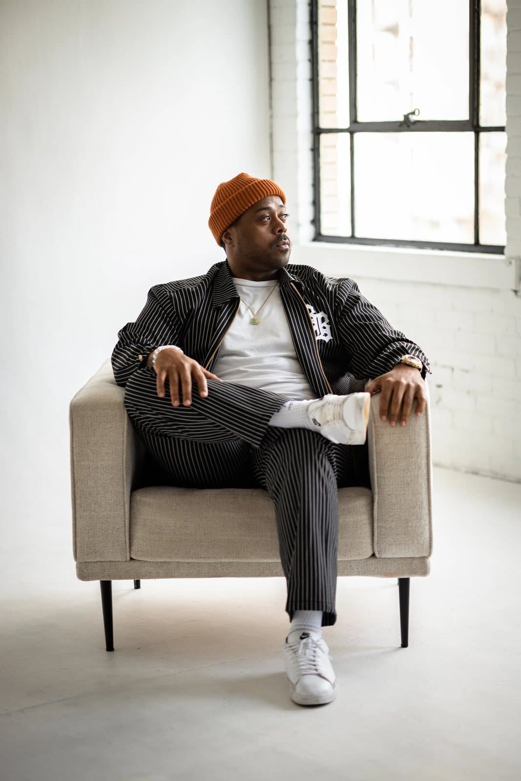 A man in an orange beanie and pinstripe suit sits in an armchair beside a bright loft window.