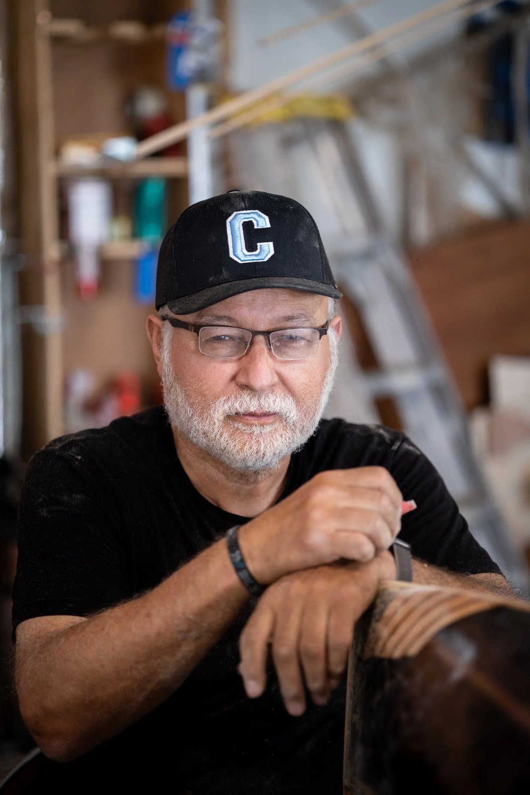 A gray-bearded man in glasses and a C cap leans on a wooden piece in his workshop.