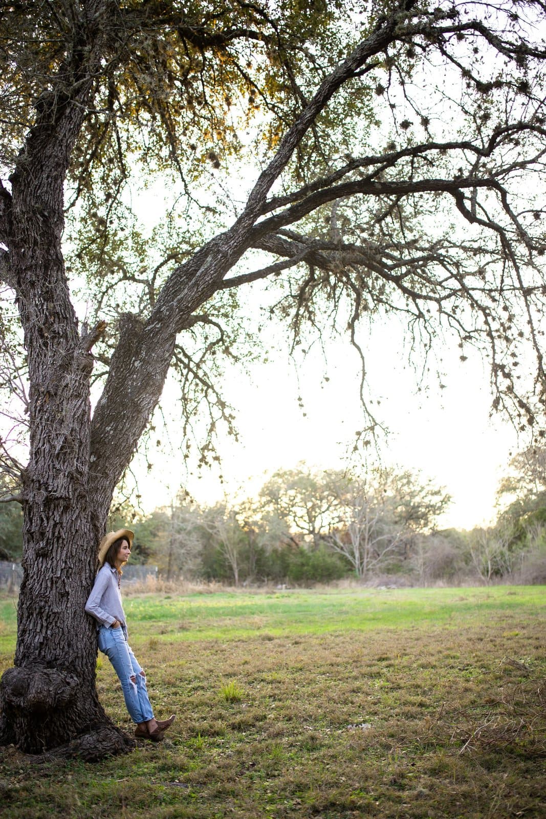 A woman in a hat leans against a large oak tree at the edge of a grassy field.