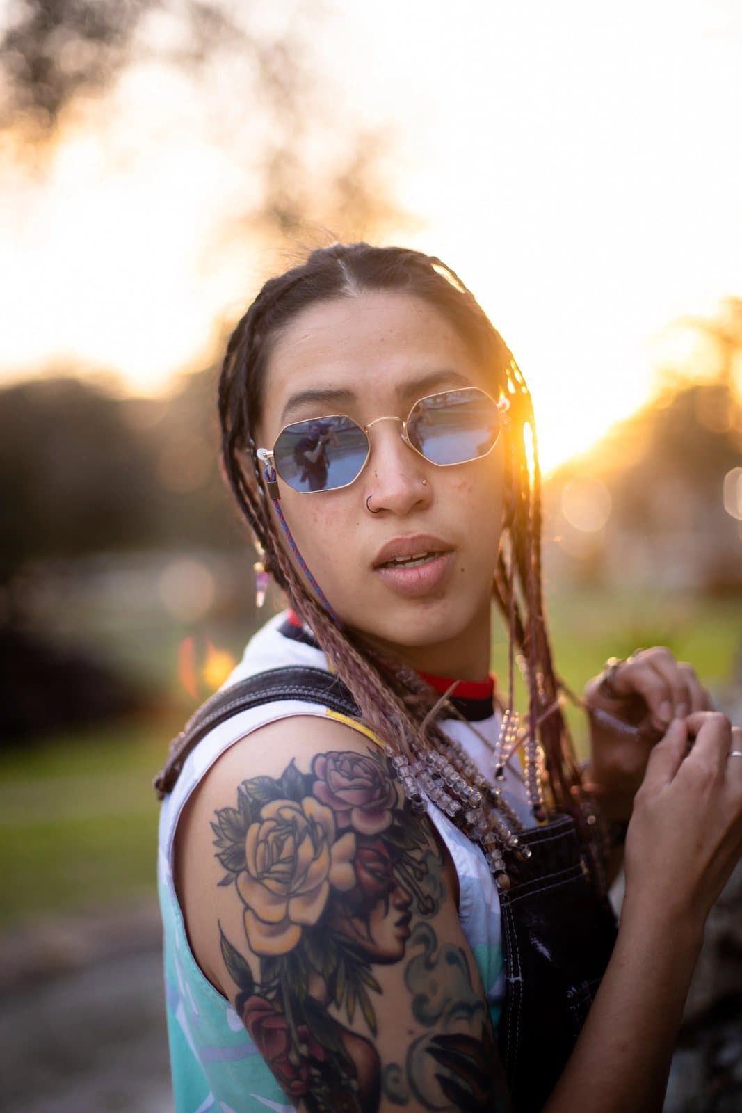 A woman with braids and blue sunglasses shows her rose tattoo in golden backlight.