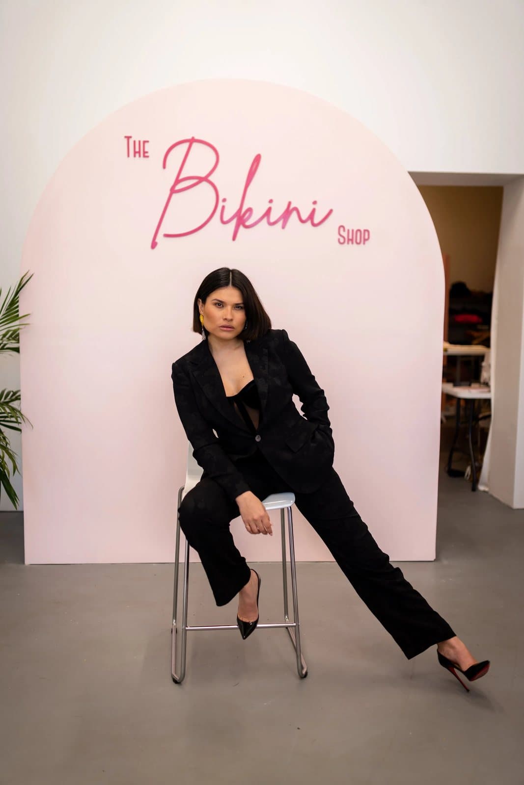 A woman in a black suit sits on a stool before a pink arch reading The Bikini Shop.