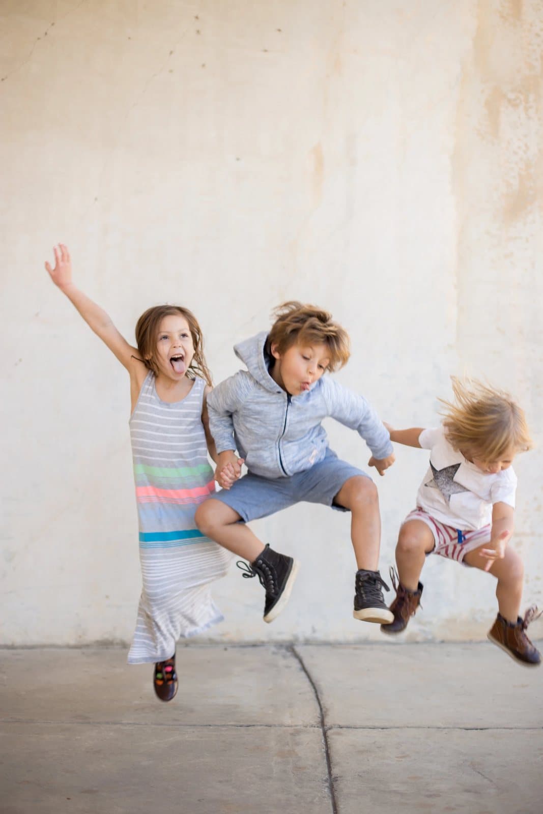 Three young children leap into the air, laughing, in front of a pale concrete wall.