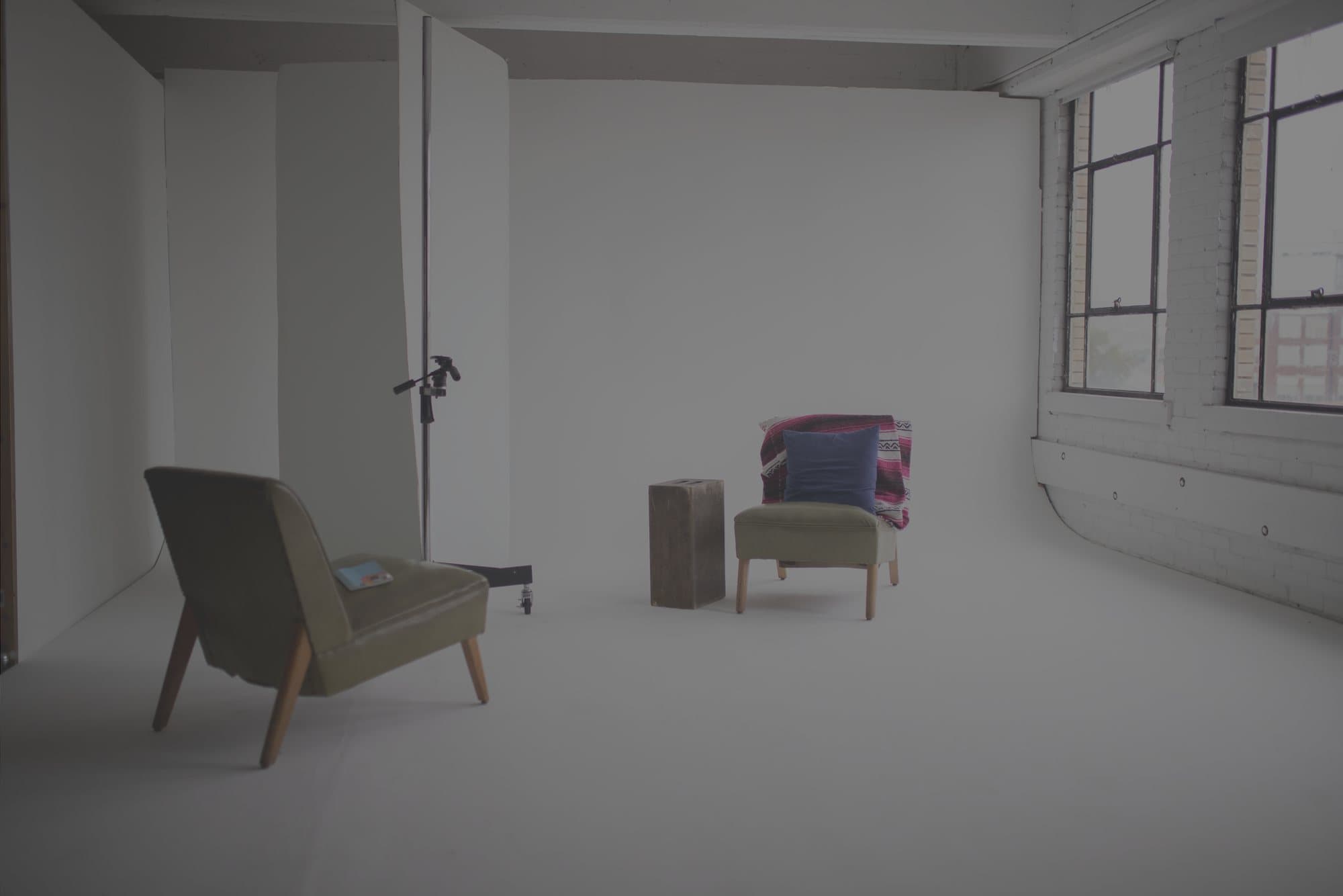 An empty white photo studio in a brick loft with two armchairs and a small side table.