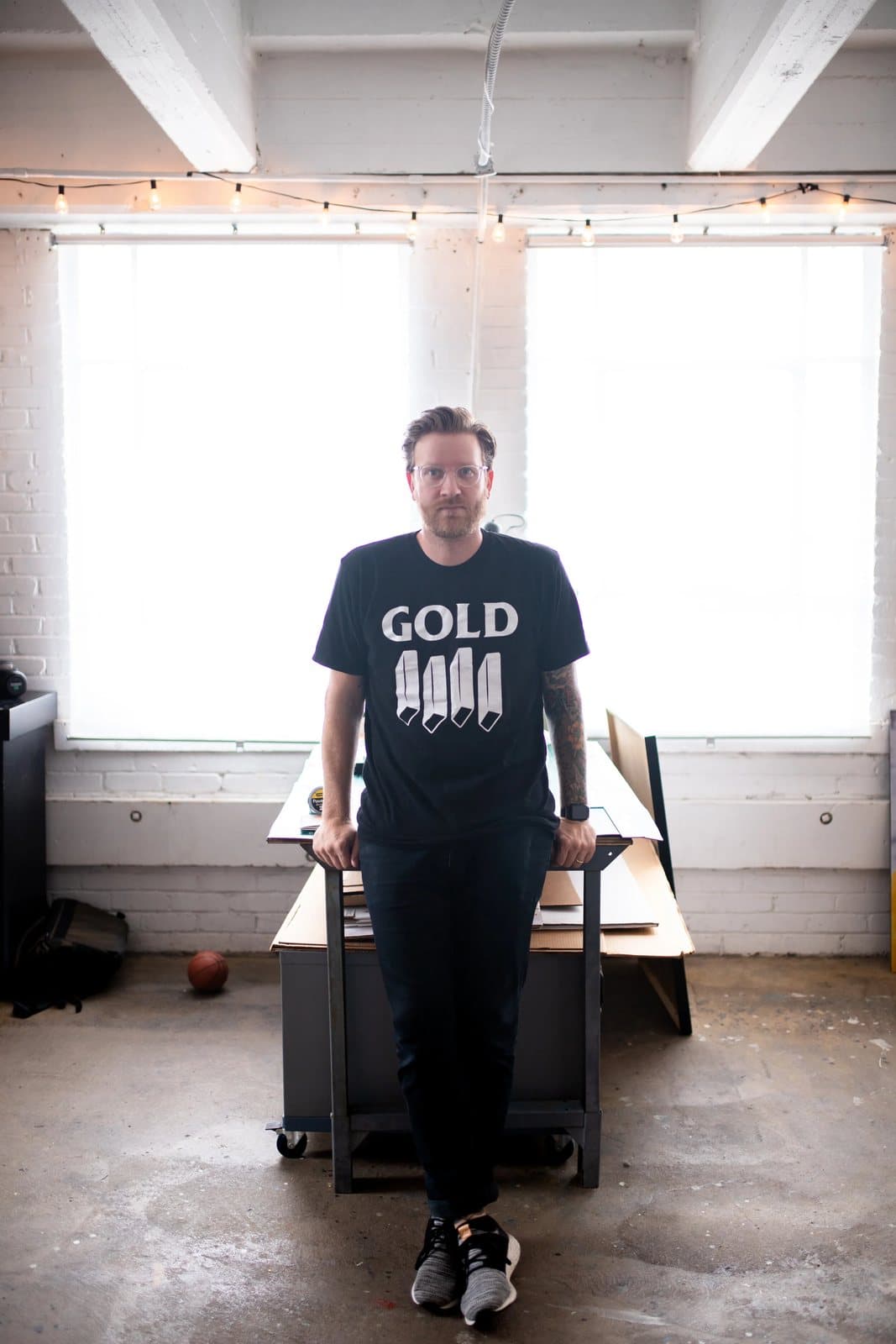A man in a GOLD t-shirt leans on a desk in a bright industrial loft strung with lights.
