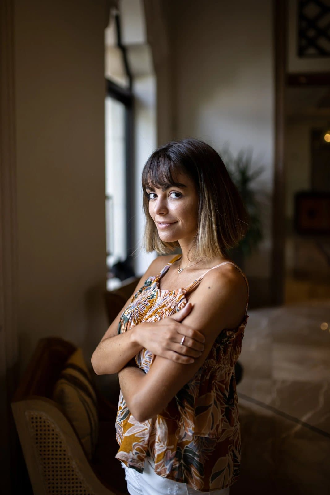 A woman with a bob and floral top stands with arms crossed by a sunlit window.