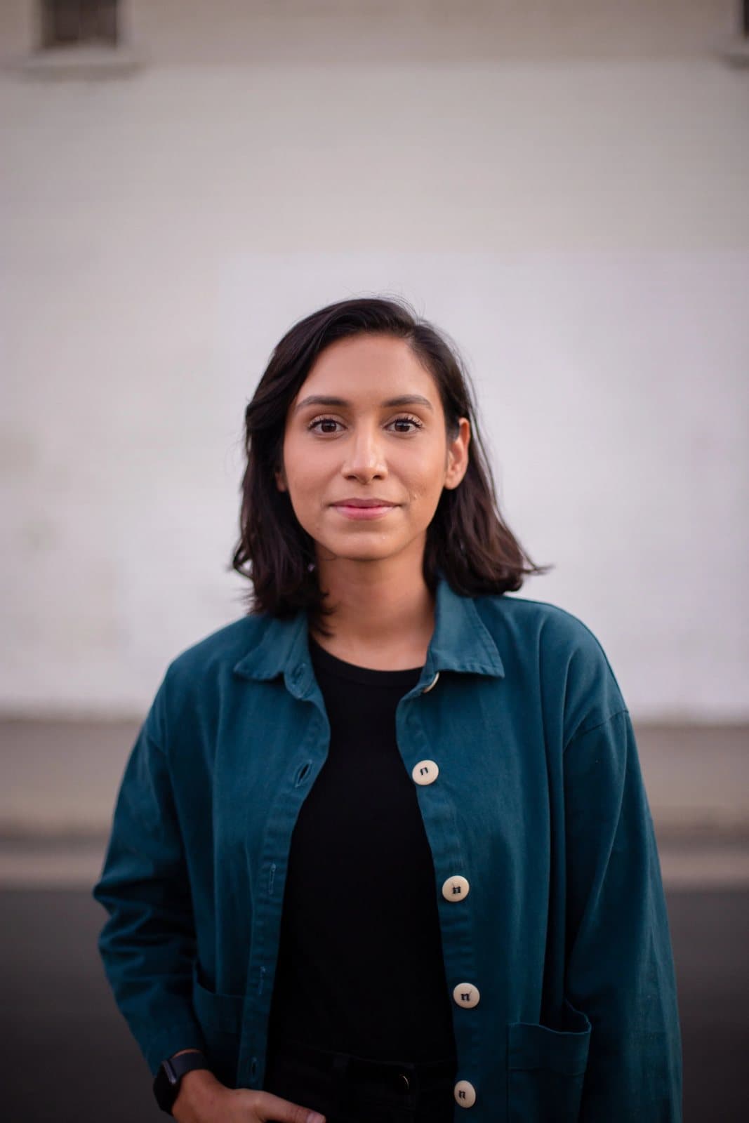 A woman in a teal jacket stands calmly against a pale wall, a natural-light portrait.