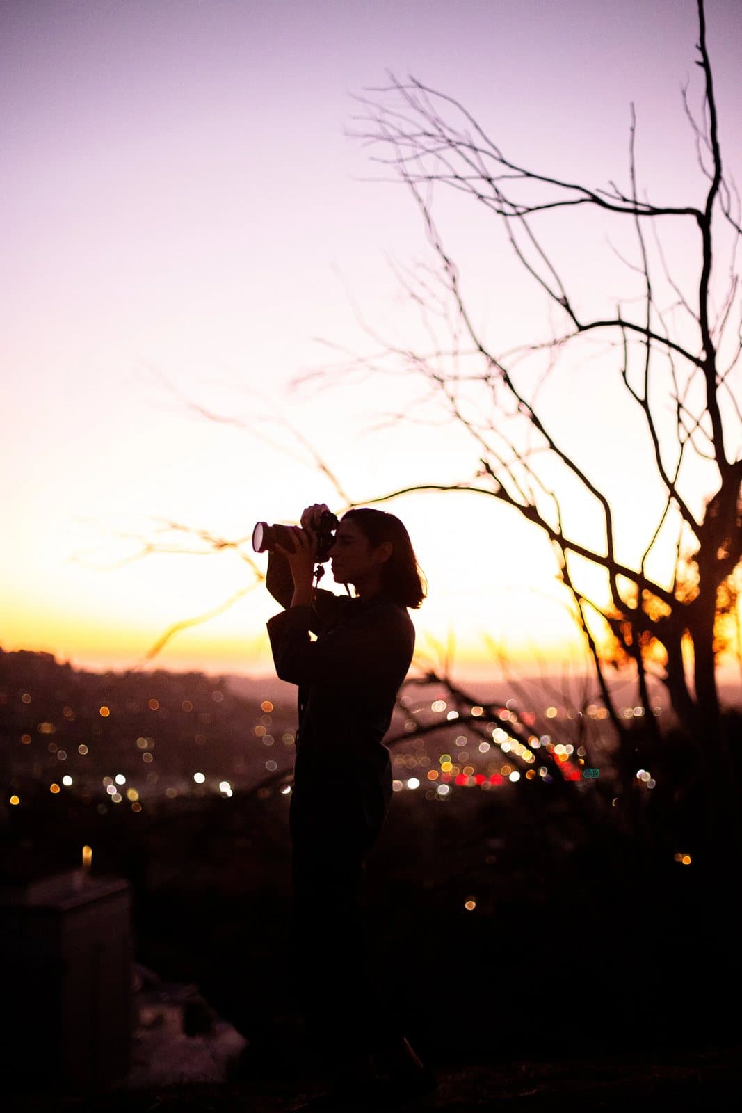 A woman silhouetted against a purple sunset raises a camera above twinkling city lights.