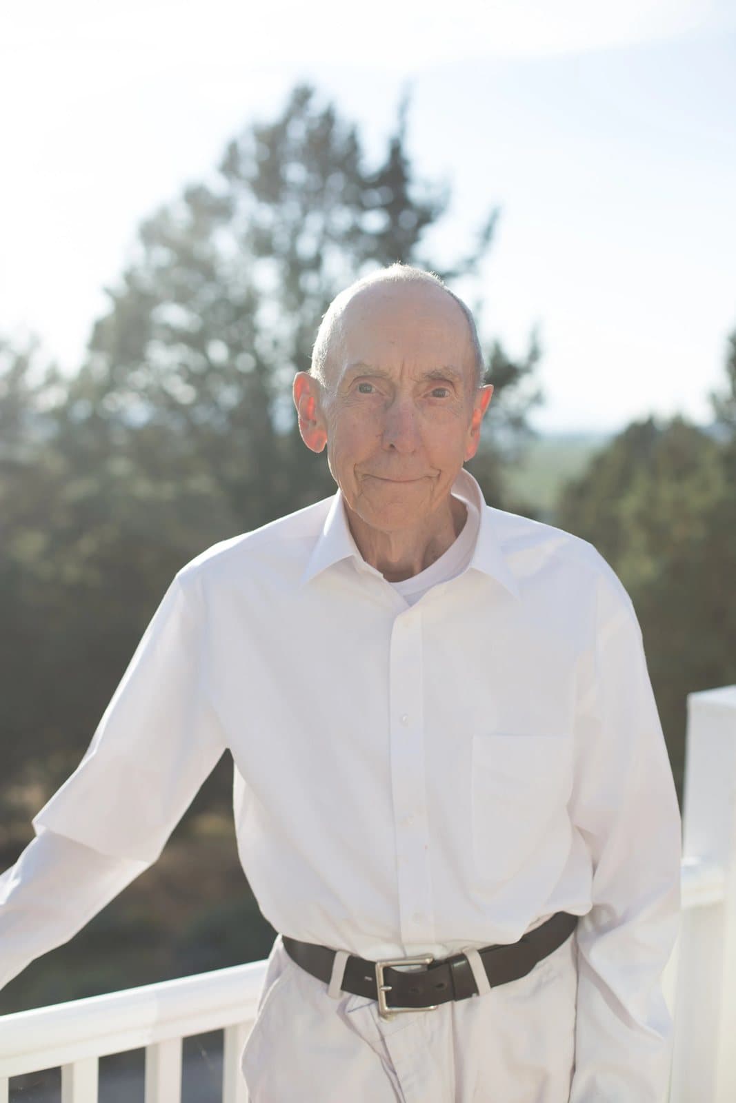 An older man in a crisp white shirt leans on a white porch railing, trees behind him.