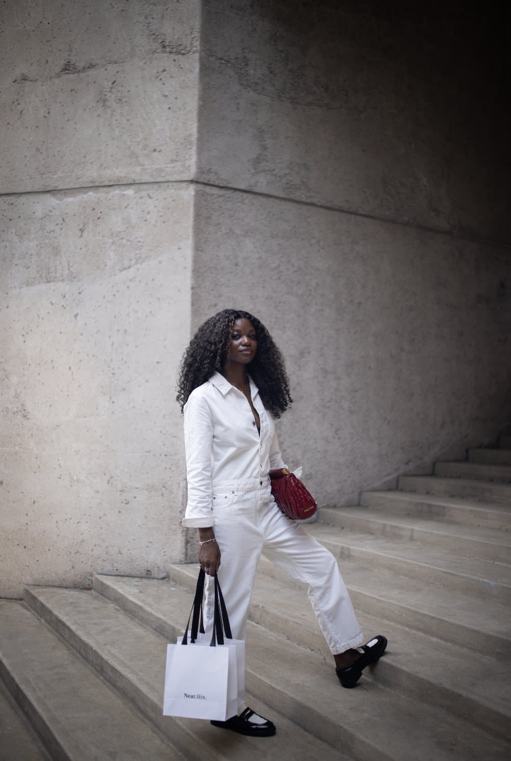 A woman in all white climbs concrete steps carrying a shopping bag and a red purse.