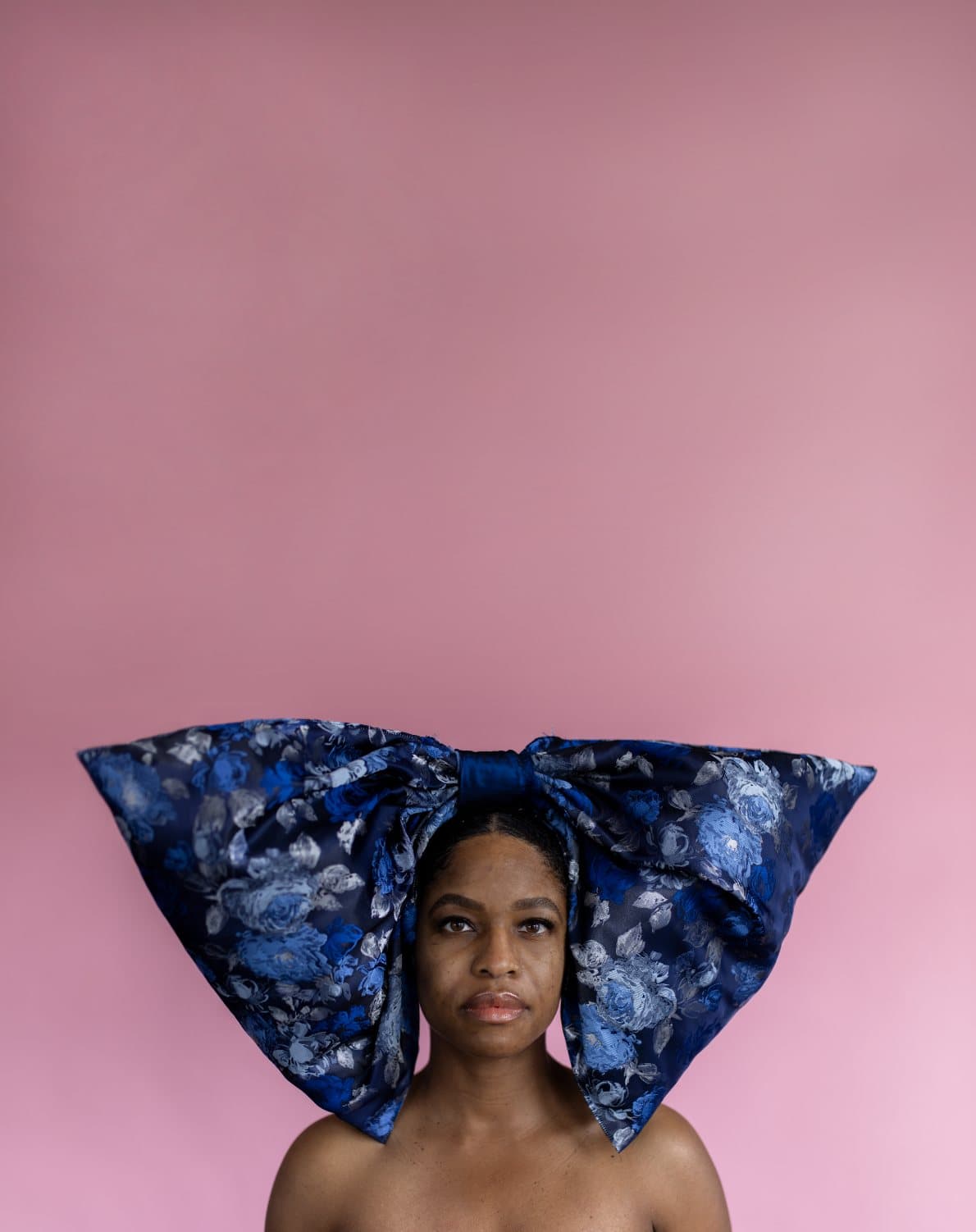 A woman wears an oversized blue floral bow on her head against a pink backdrop.