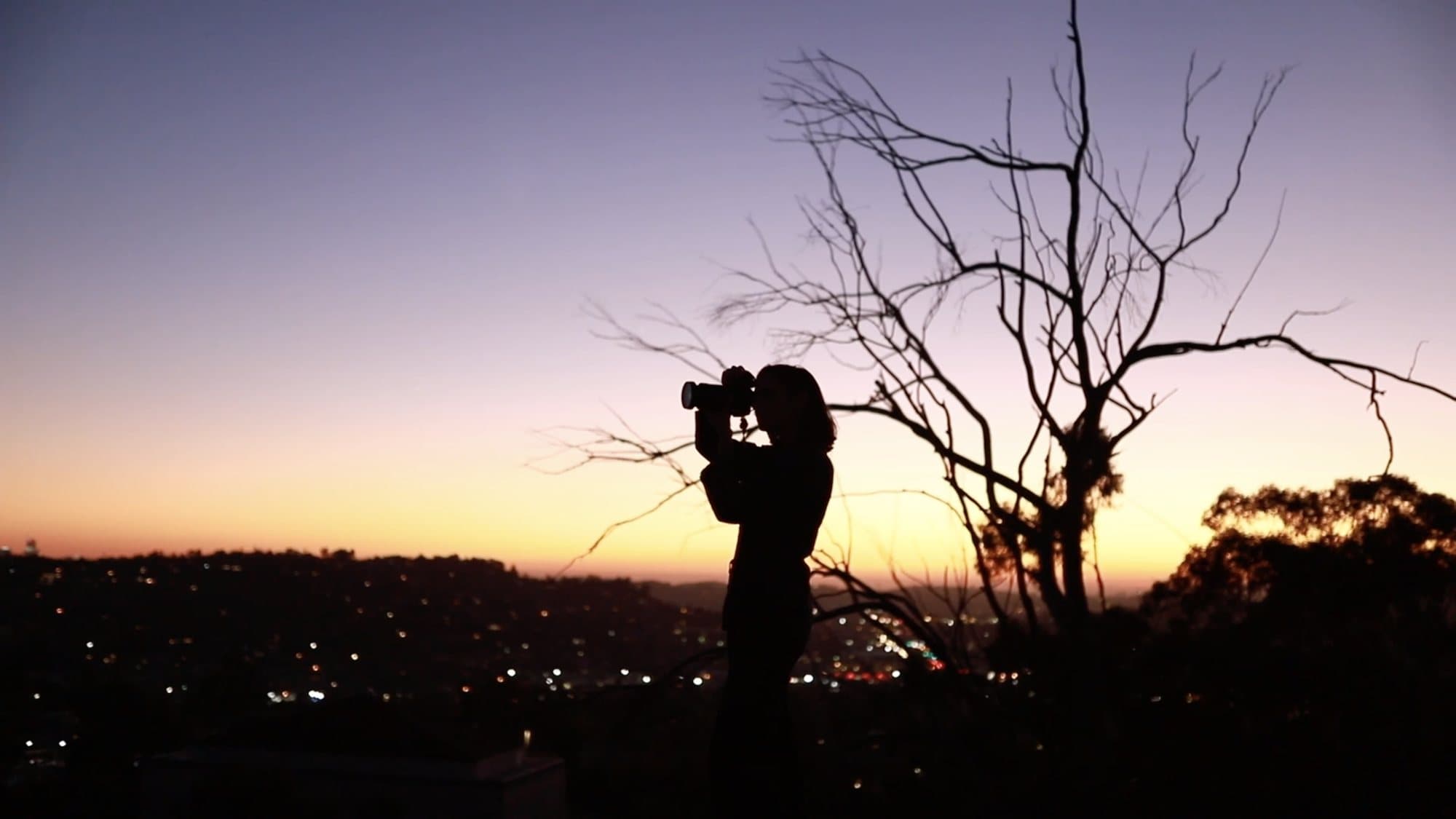 Video teaser still of a woman silhouetted with a camera against a sunset over city lights.