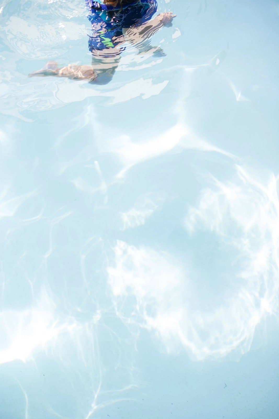 A child in a patterned swimsuit swims across sunlit, rippling pool water.