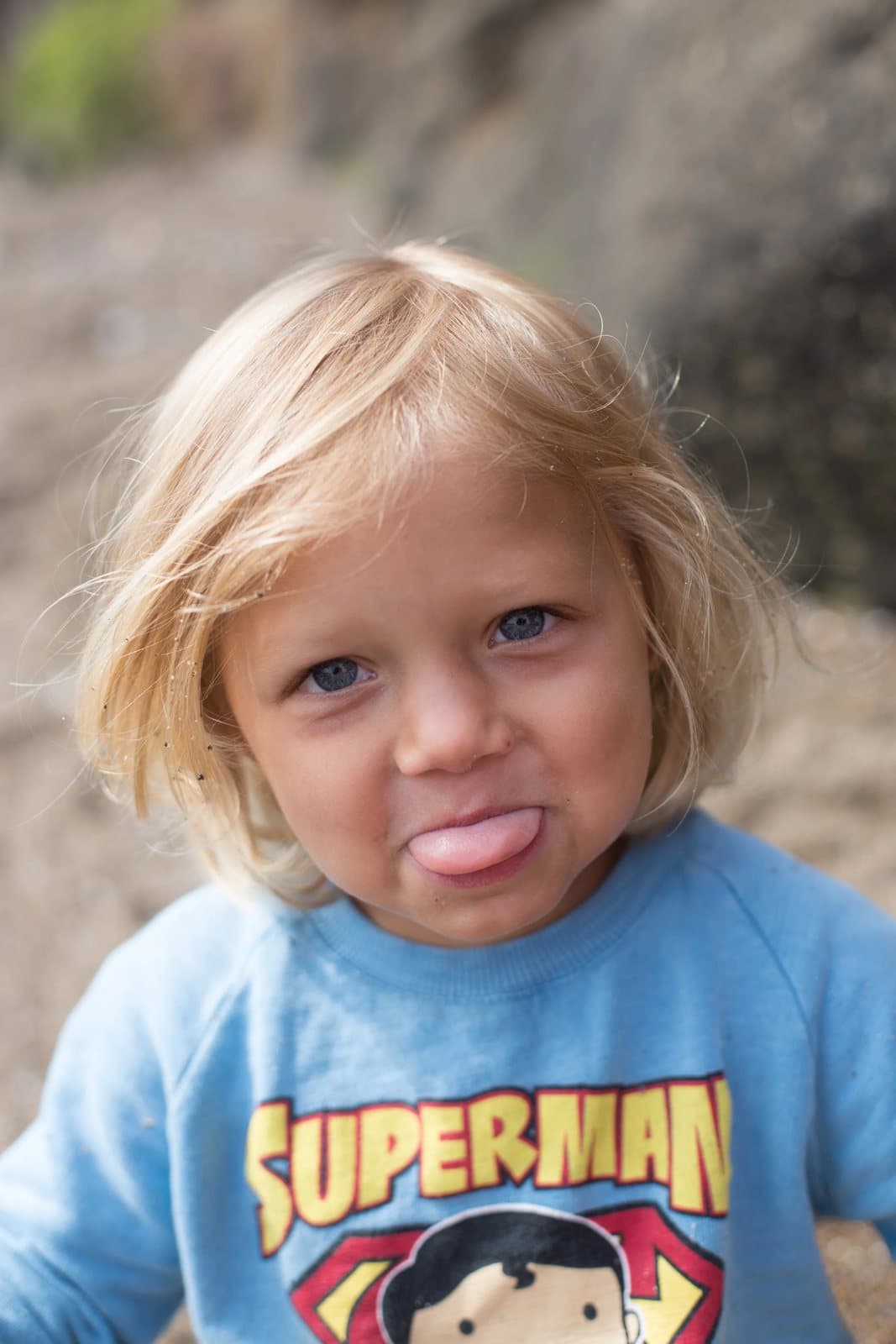 A blond toddler in a Superman sweatshirt sticks out his tongue at the camera.