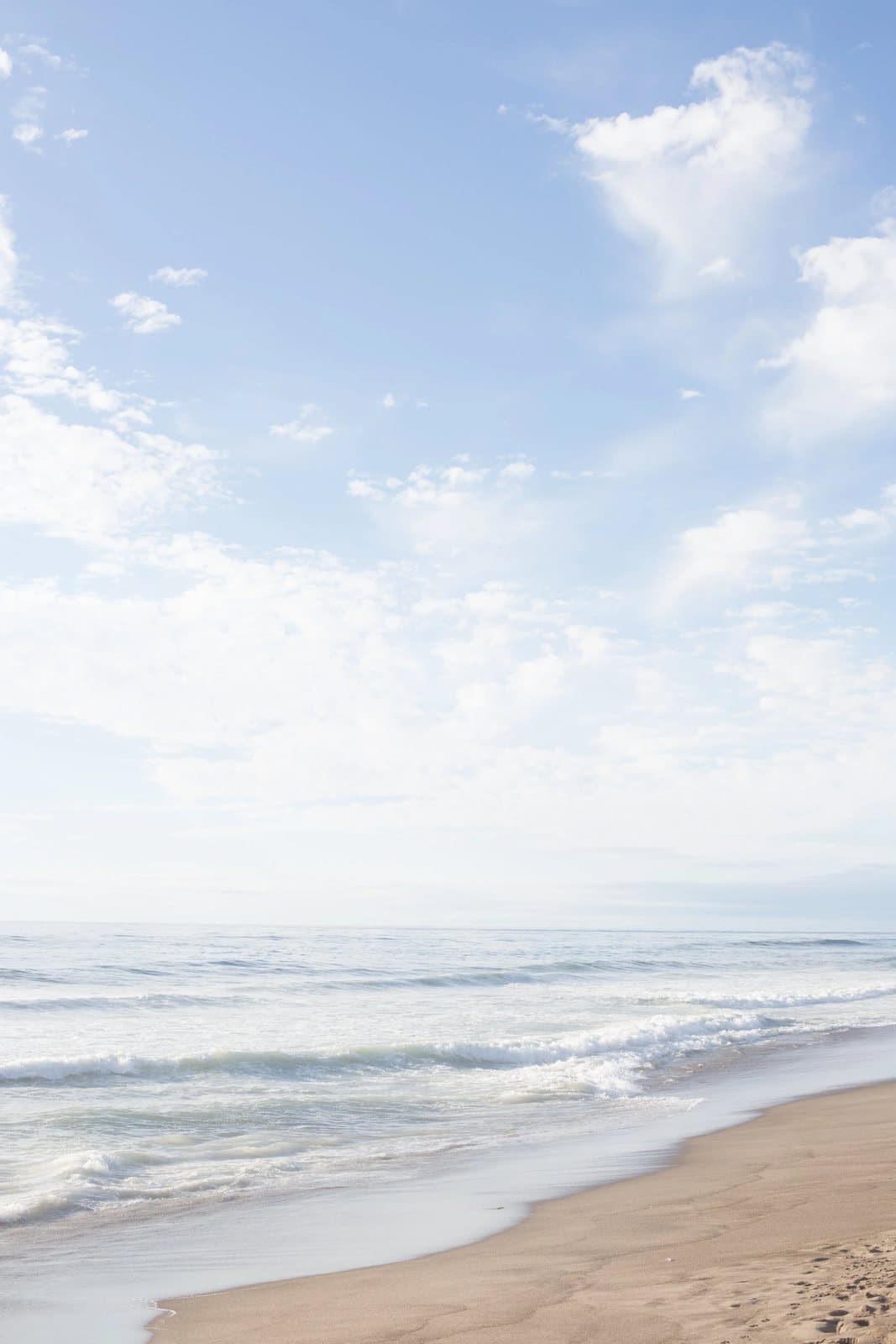 Soft clouds in a pale blue sky above gentle waves on a sandy beach.