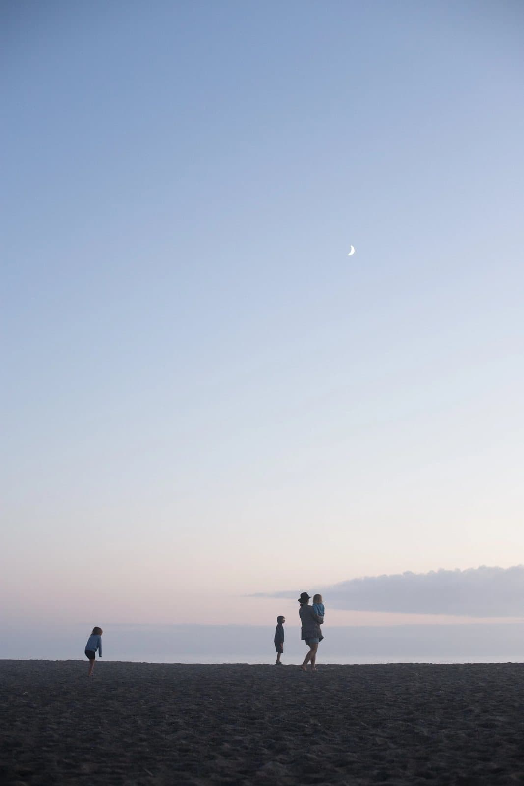 Small figures walk a dark beach at dusk beneath a crescent moon.