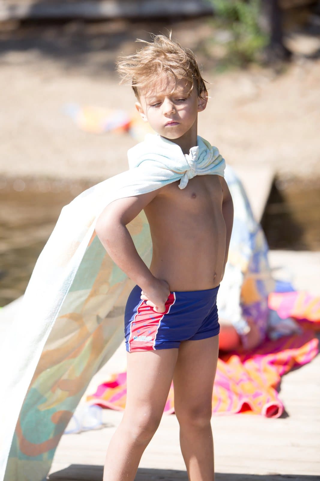 A boy wearing a towel as a cape stands hands on hips on a sunny dock.