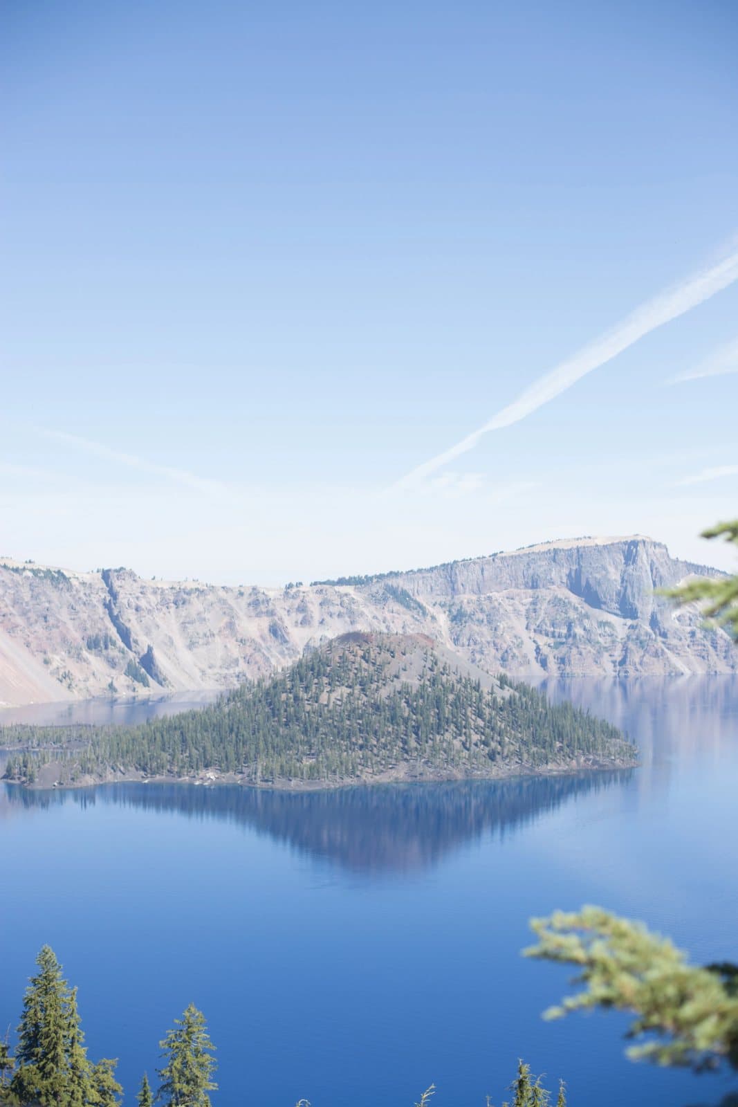 Crater Lake and Wizard Island beneath a clear blue sky, framed by pine branches.