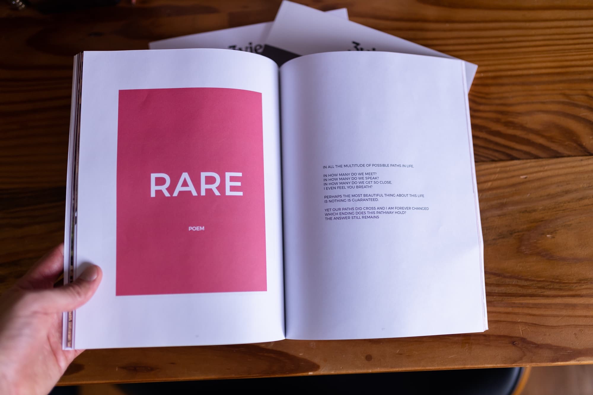 Hands holding the 3vies magazine open to a spread with a pink panel titled Rare, a poem, on a wooden table.